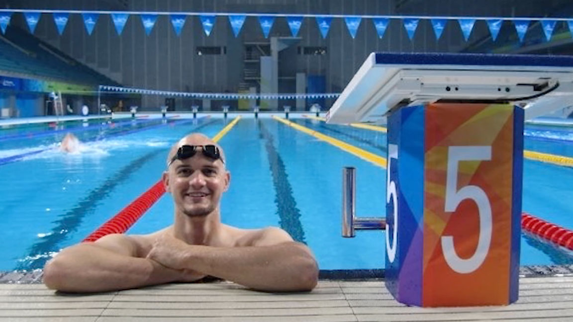 Dave Denniston in an indoor swimming pool. Dave is hanging off the edge of the pool, with googles on top of head and smiling at the camera
